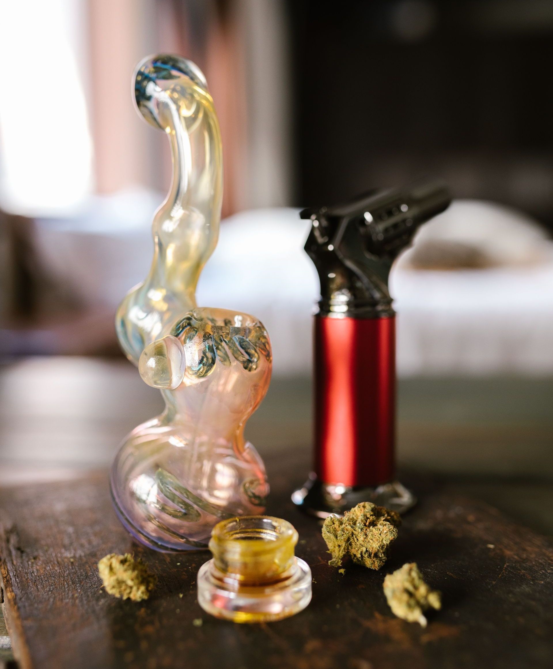 Artistic glass bubbler with cannabis buds and torch lighter on a wooden table.